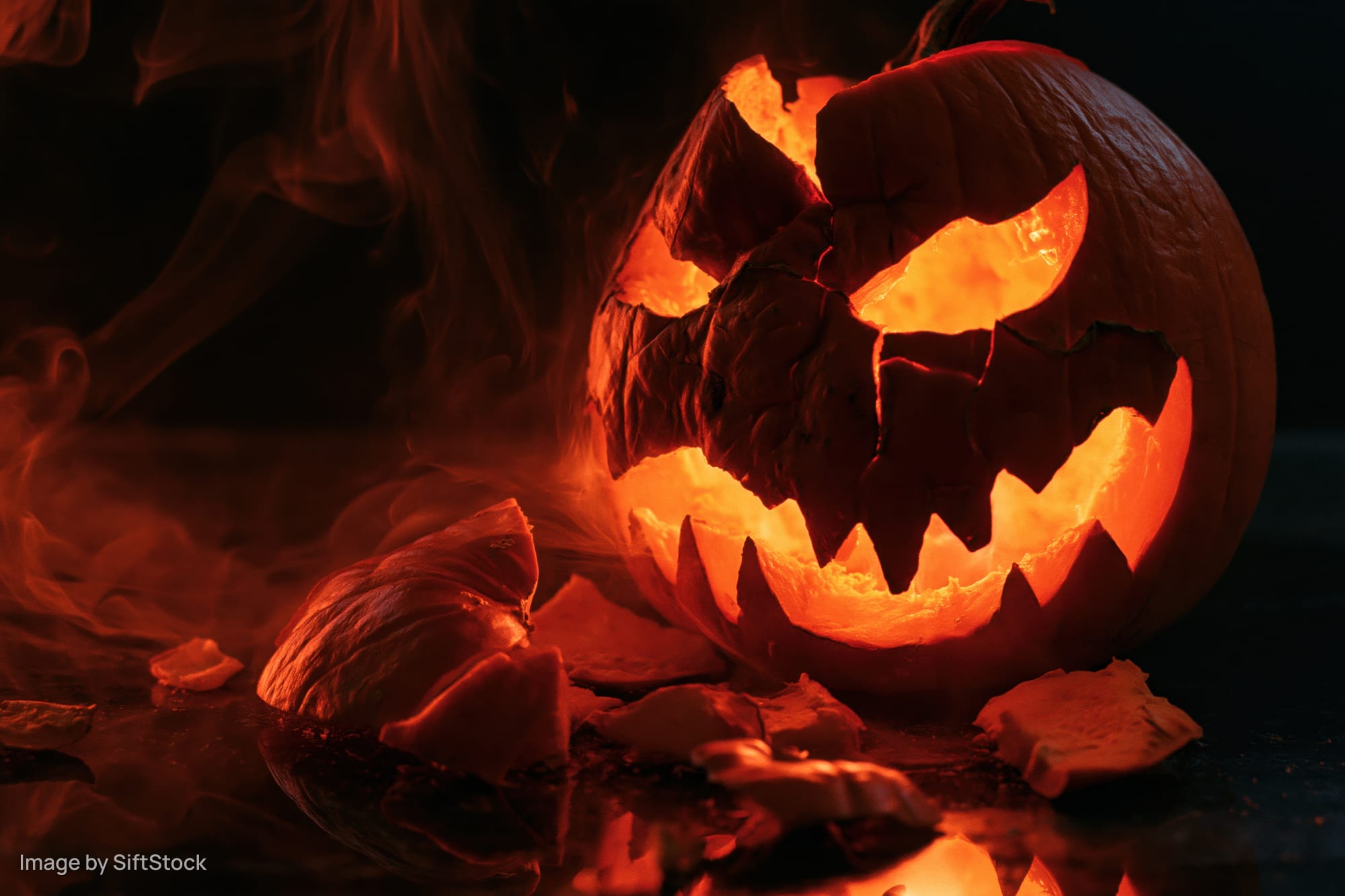 Cracked jack-o-lantern glowing with orange light on dark wet surface surrounded by broken pieces and smoke