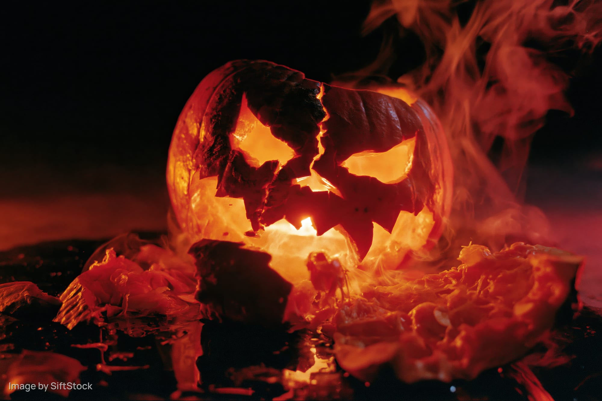 Cracked jack-o-lantern glowing with smoke rising from broken pieces on dark ground
