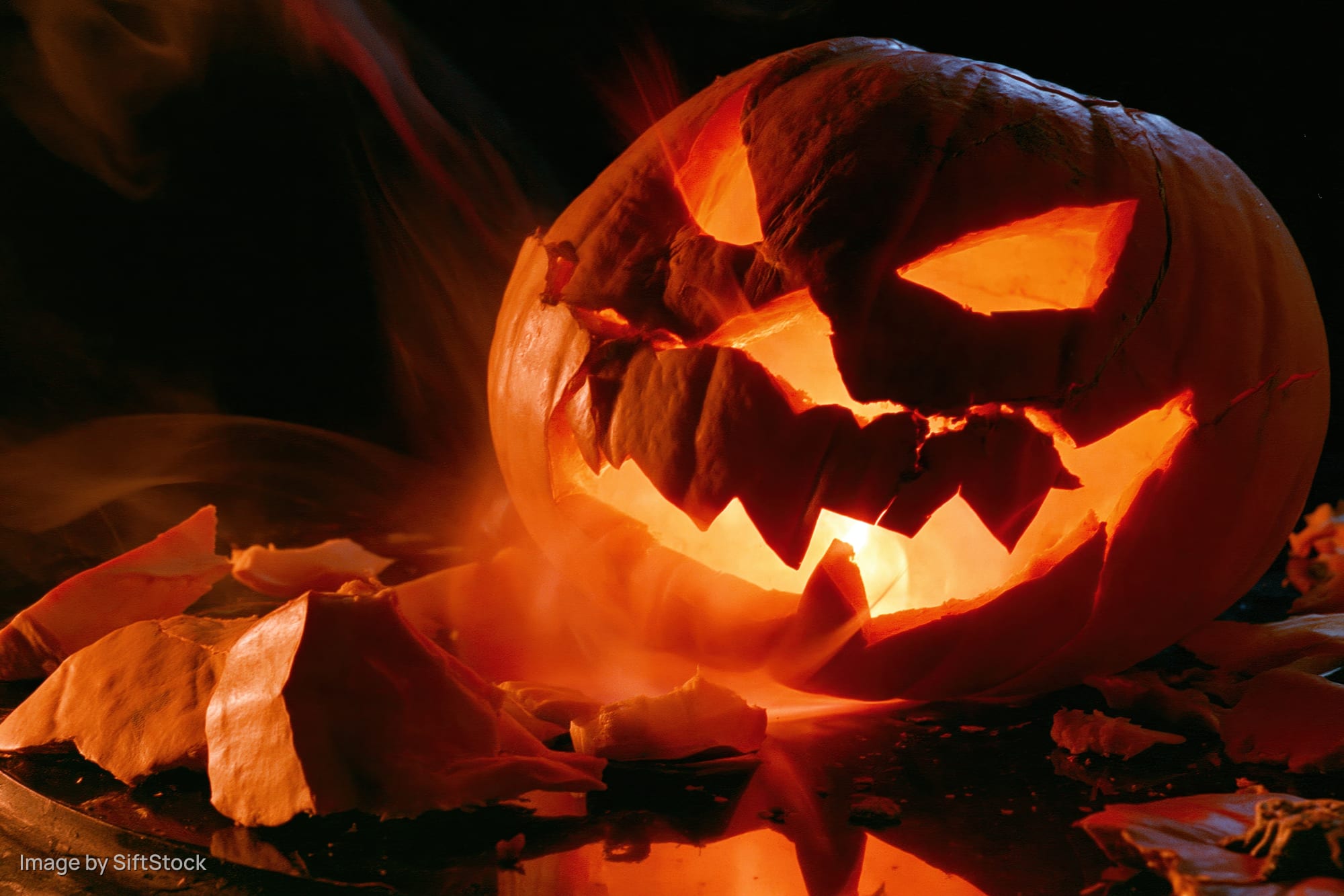 Cracked jack-o-lantern glowing with candlelight on dark surface surrounded by broken pieces