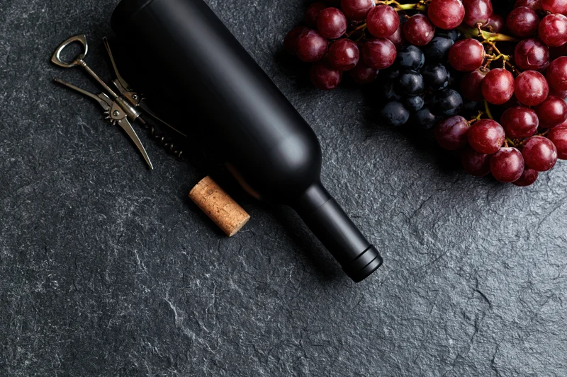 Dark wine bottle lying flat beside cork and corkscrew with grapes on slate surface