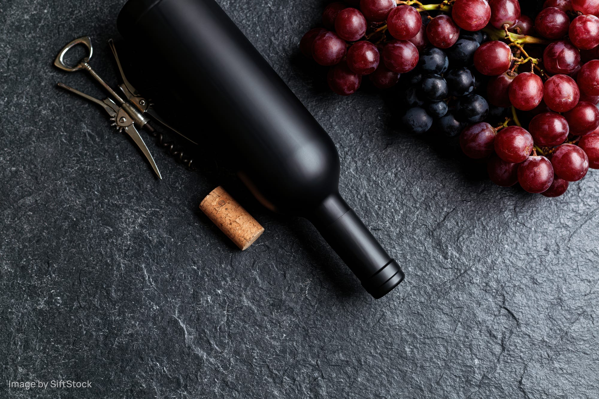 Dark wine bottle lying flat beside cork and corkscrew with grapes on slate surface