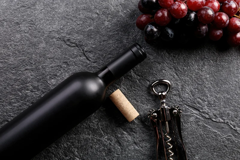 Dark wine bottle lying flat beside cork and corkscrew with grapes on slate surface