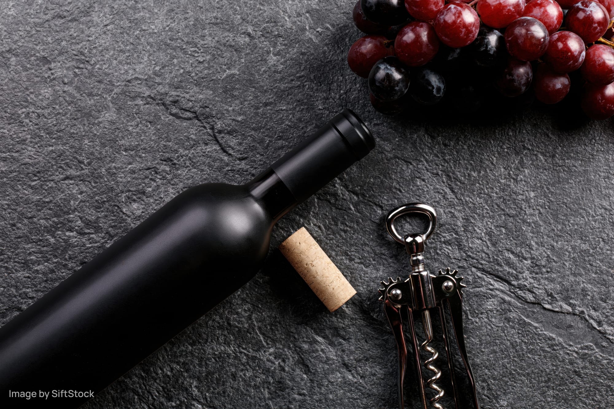 Dark wine bottle lying flat beside cork and corkscrew with grapes on slate surface