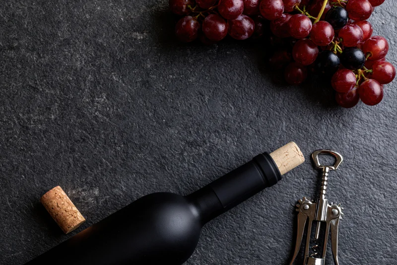 Dark wine bottle lying flat beside cork and corkscrew with grapes on slate surface