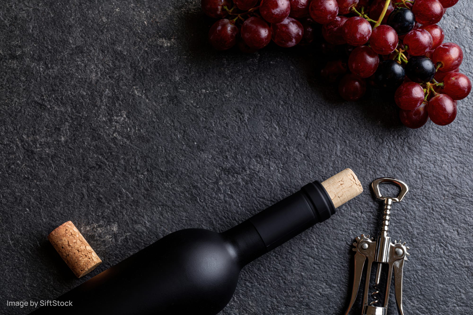 Dark wine bottle lying flat beside cork and corkscrew with grapes on slate surface