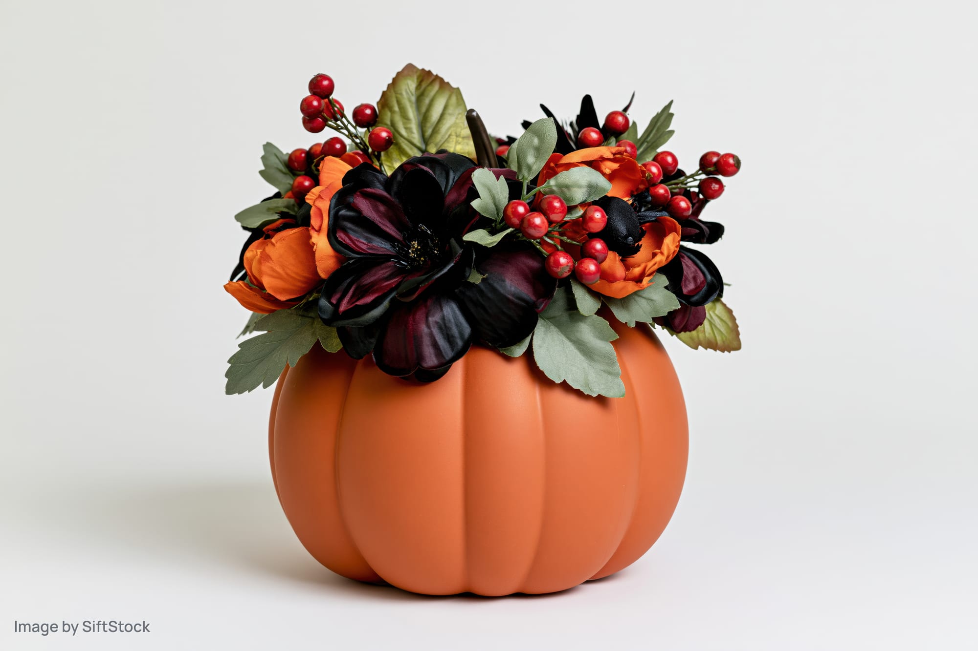 Ceramic pumpkin centerpiece topped with artificial black and orange flowers and red berries on white background
