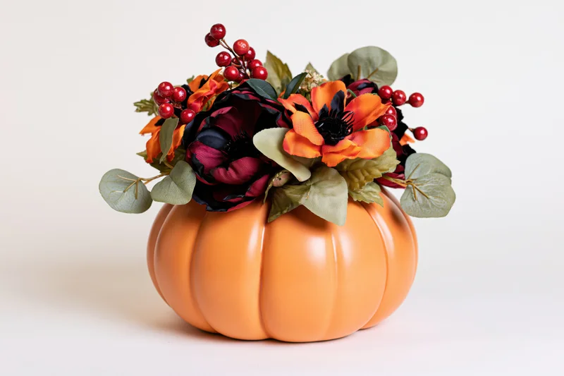 Ceramic pumpkin centerpiece topped with artificial flowers and berries on white background