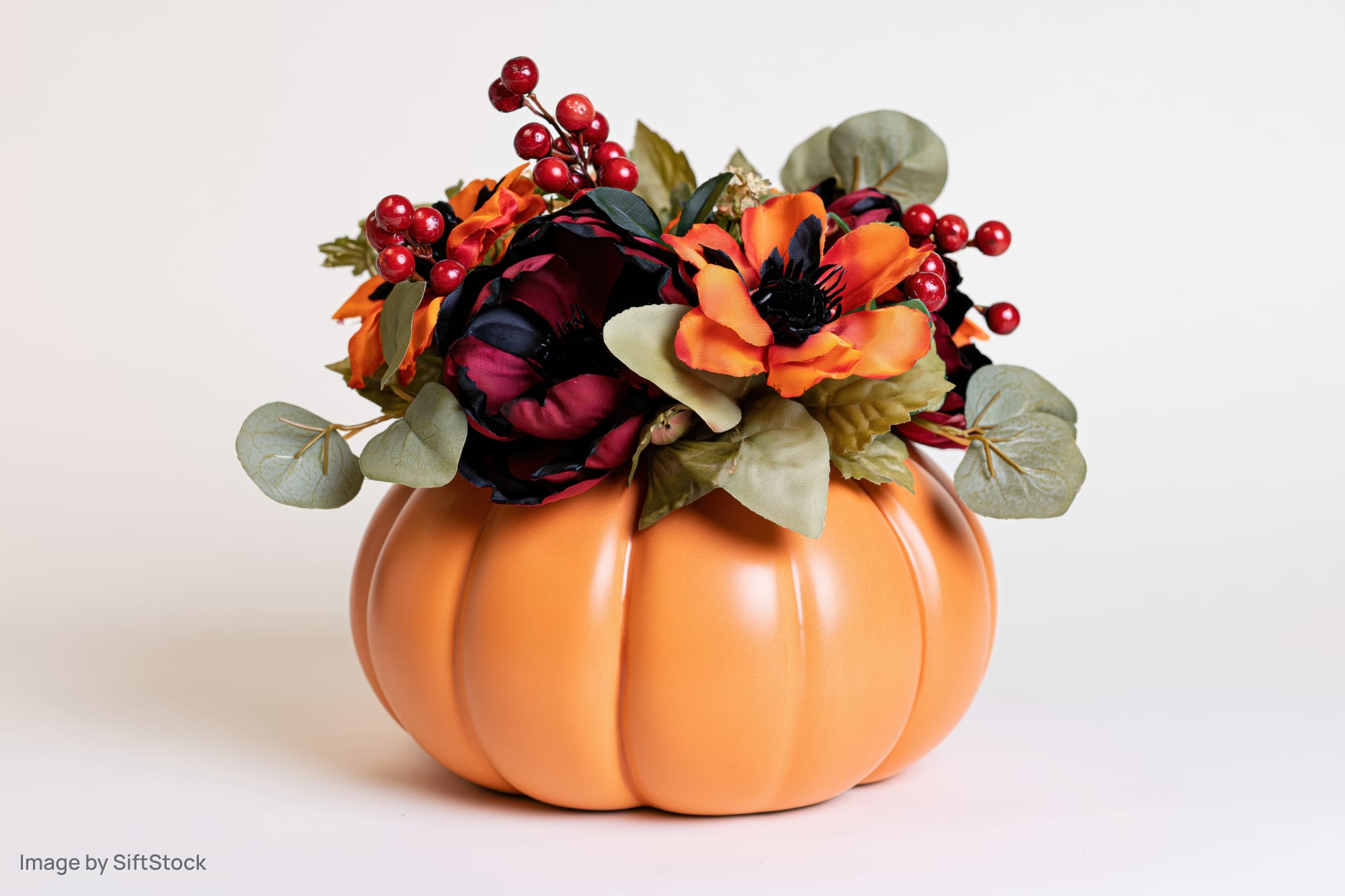 Ceramic pumpkin centerpiece topped with artificial flowers and berries on white background