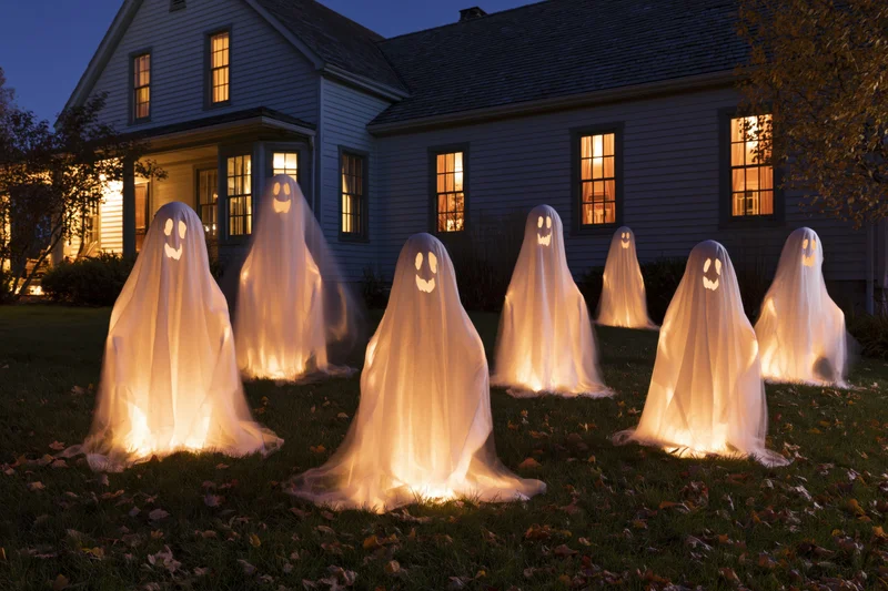 Glowing white ghost decorations arranged on a front lawn at night in front of a house with lit windows