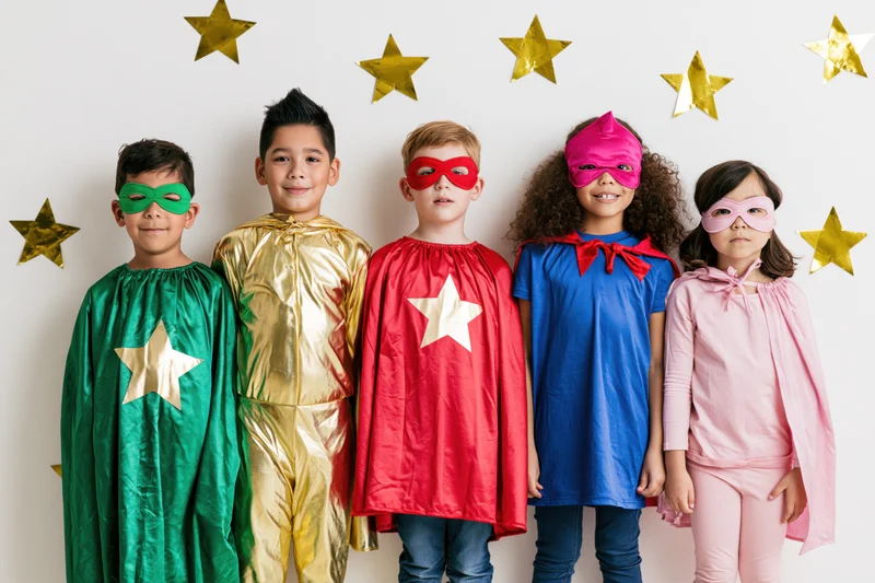 Five children in colorful superhero costumes posing together against a white wall with gold stars