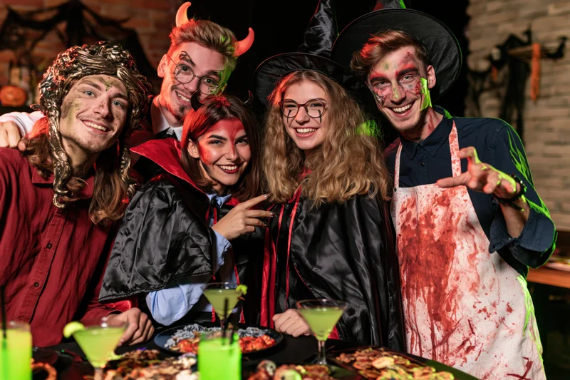 Five young adults in halloween costumes posing together at a party table with drinks and snacks