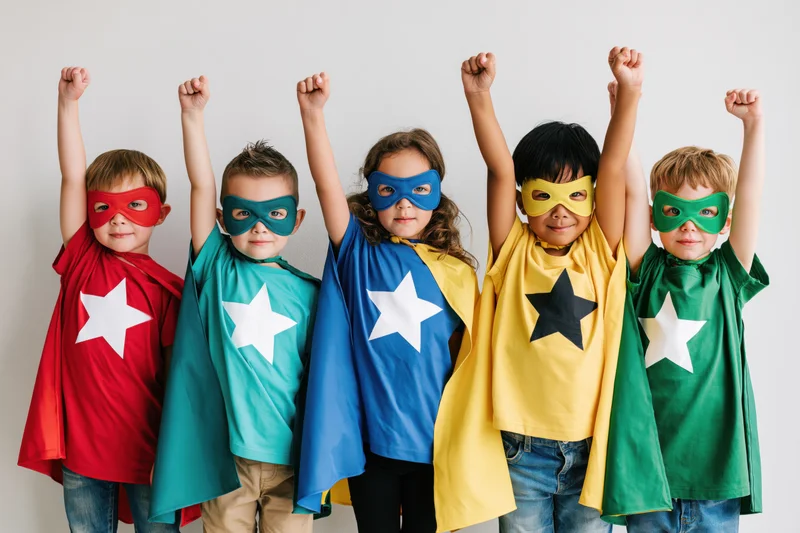 Five children in superhero costumes with capes and masks raising fists in a line against a plain background
