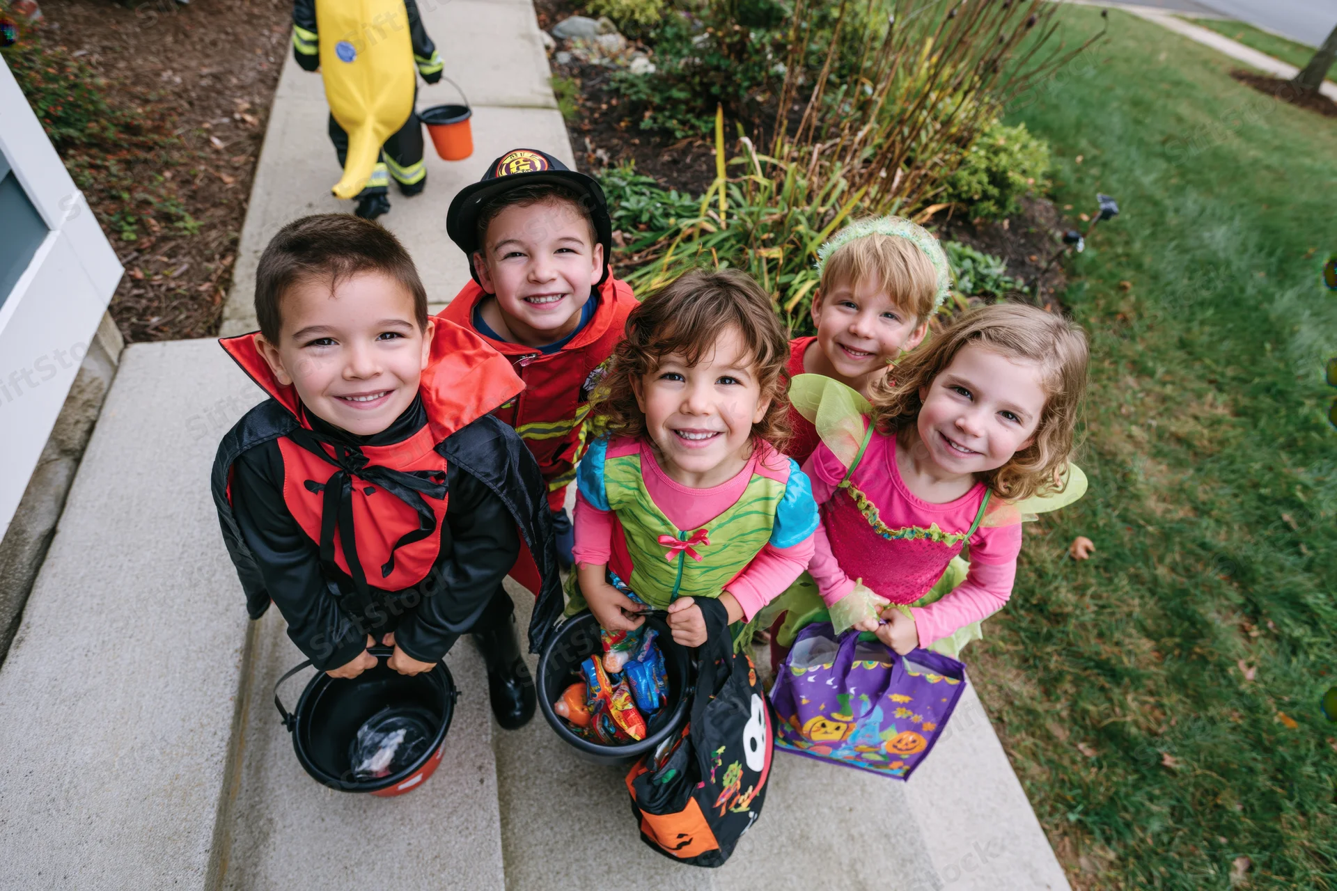 Five young children in halloween costumes smiling while holding candy buckets on front steps