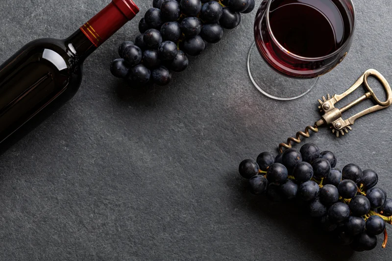 Dark wine bottle, glass of red wine, grapes and corkscrew arranged on slate surface