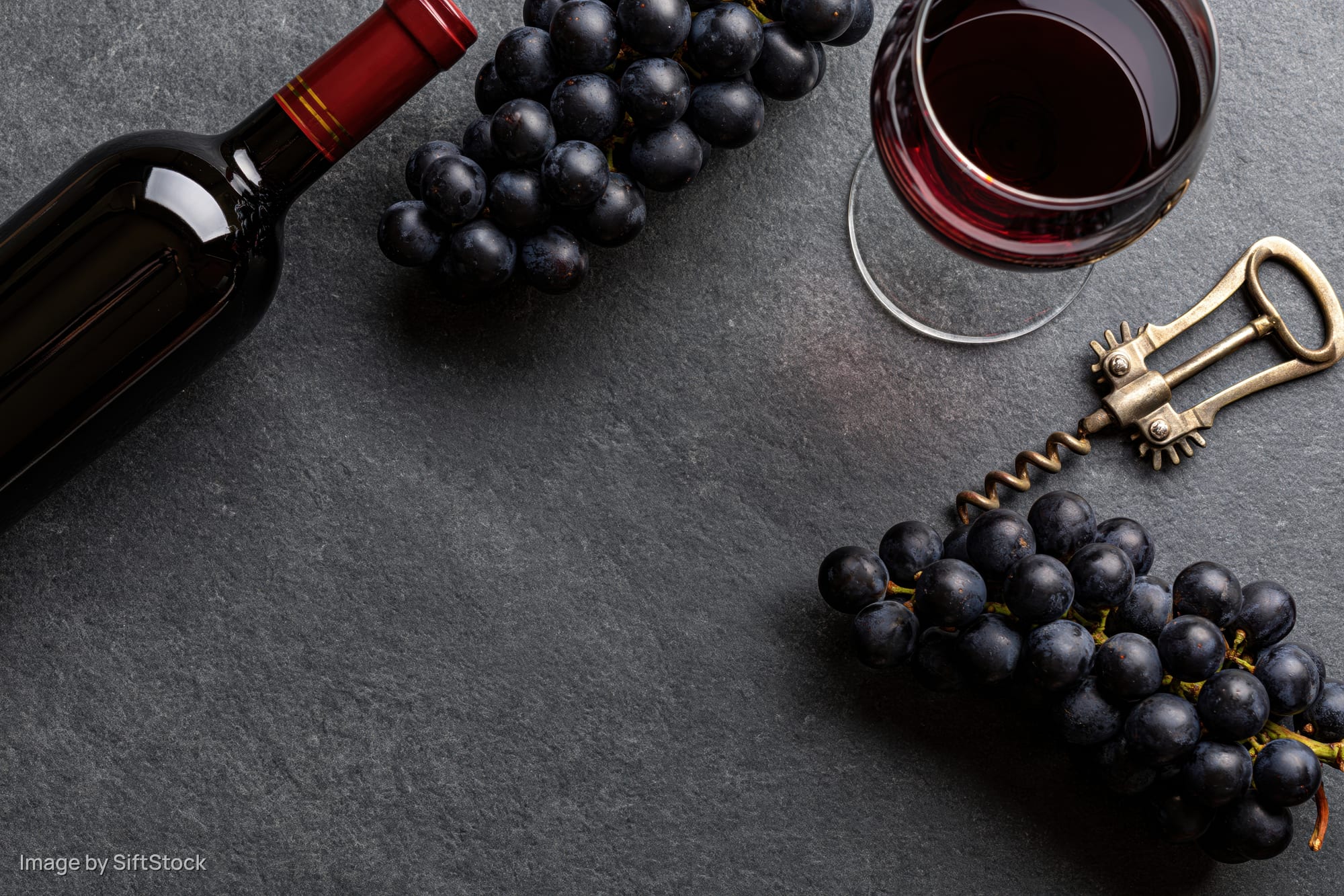 Dark wine bottle, glass of red wine, grapes and corkscrew arranged on slate surface