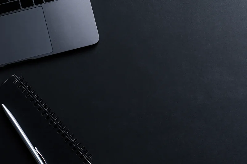 Silver laptop and notebook with pen on black desk surface, top view