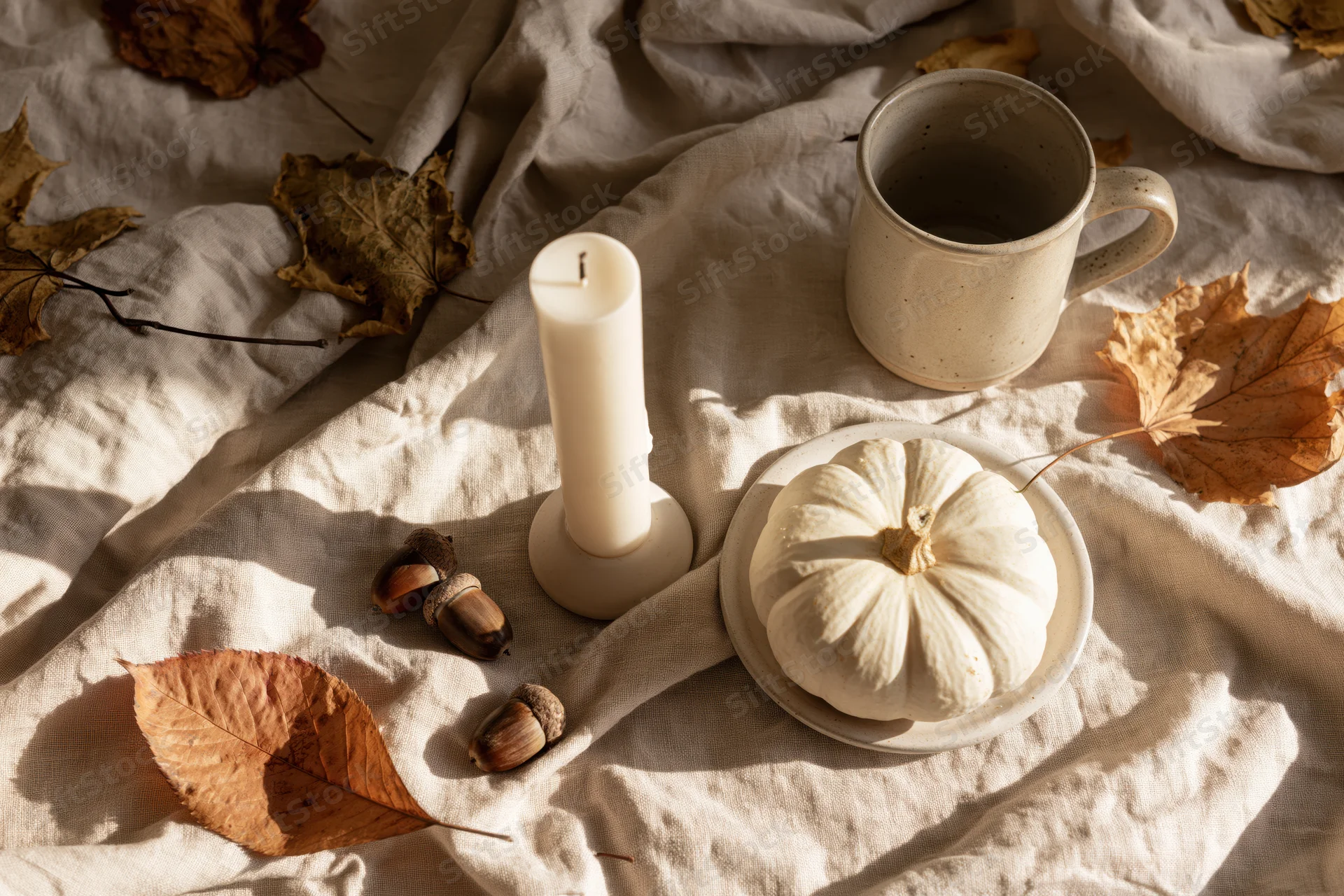 Small white pumpkin and unlit candle on linen with autumn leaves and acorns