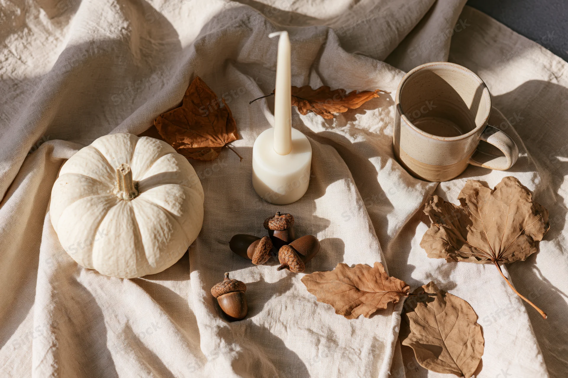 White pumpkin, candle, acorns and dried leaves arranged on linen fabric in soft sunlight