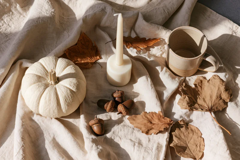 White pumpkin, candle, acorns and dried leaves arranged on linen fabric in soft sunlight