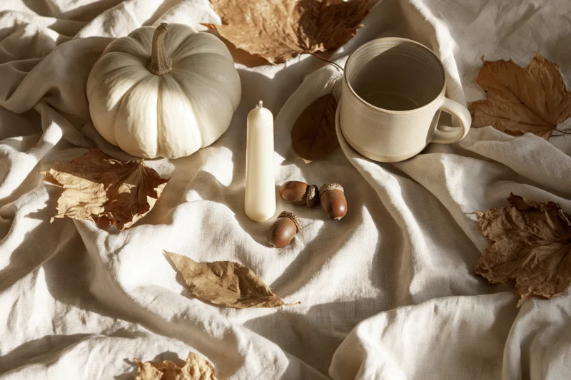White pumpkin, candle, acorns and dried leaves arranged on crumpled linen fabric