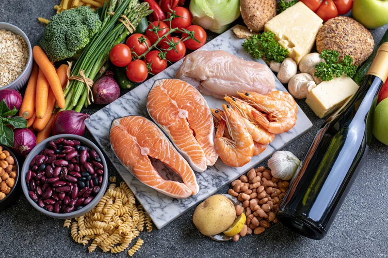 Salmon and shrimp fillets on a marble cutting board surrounded by fresh vegetables, fruits, grains, legumes, dairy, and wine in a flat lay arrangement