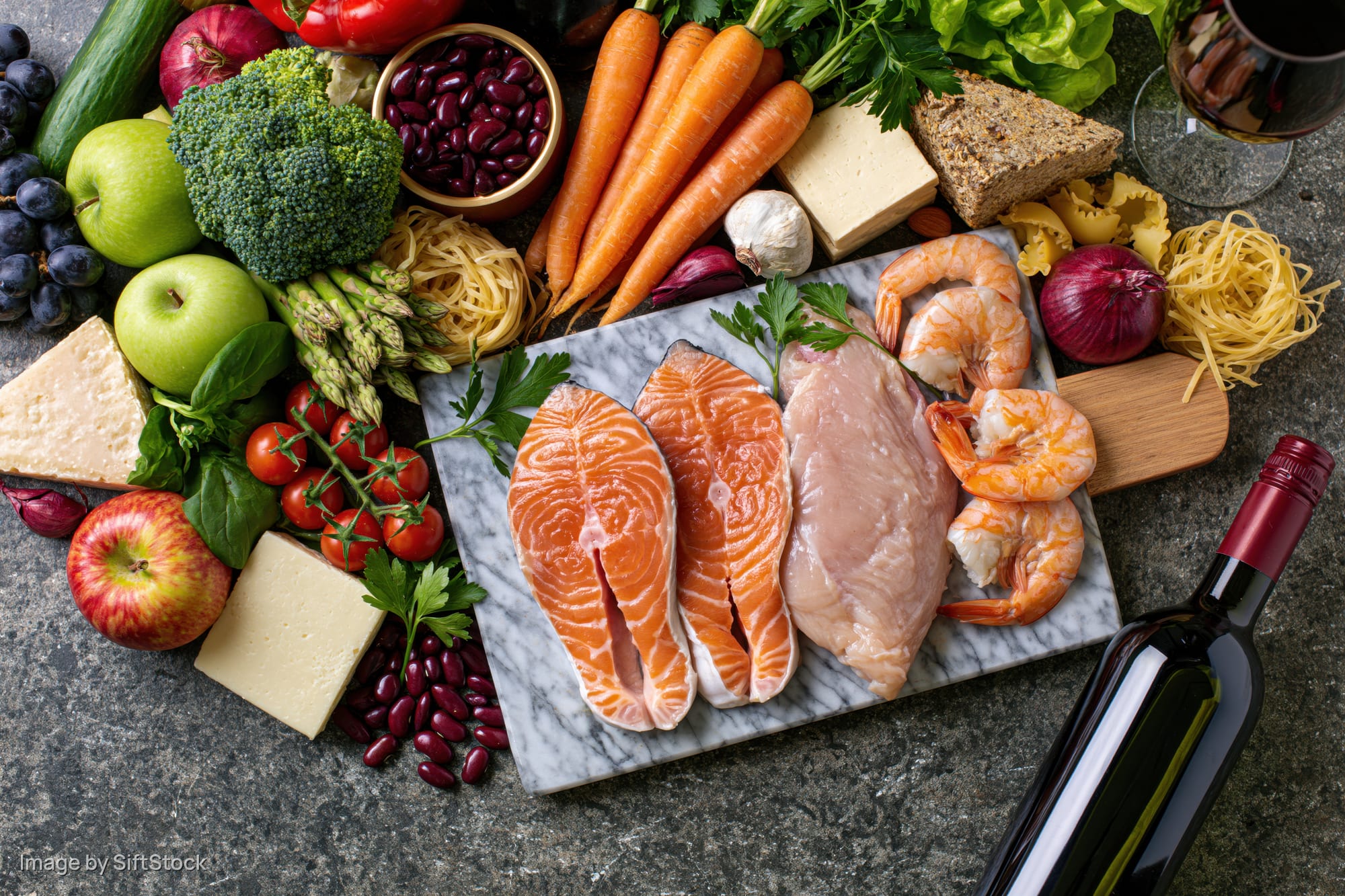 Salmon, chicken and shrimp on a marble cutting board surrounded by vegetables, fruits, cheese and wine