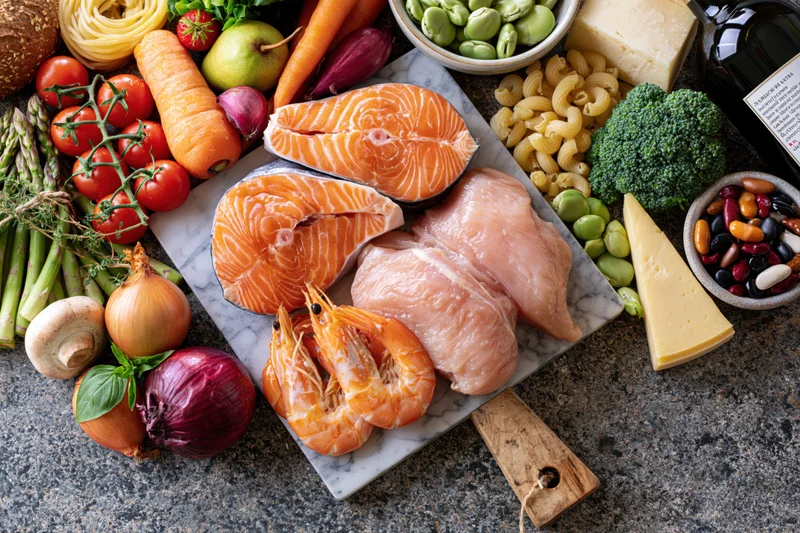 Salmon and chicken fillets with shrimp on a marble cutting board surrounded by fresh vegetables, fruits, dairy, grains, and legumes