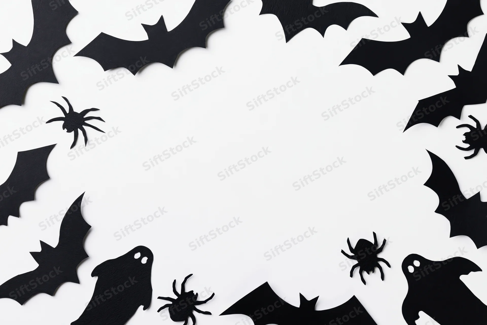 Black paper halloween cutouts of bats spiders and ghosts arranged in a frame on white background with copyspace