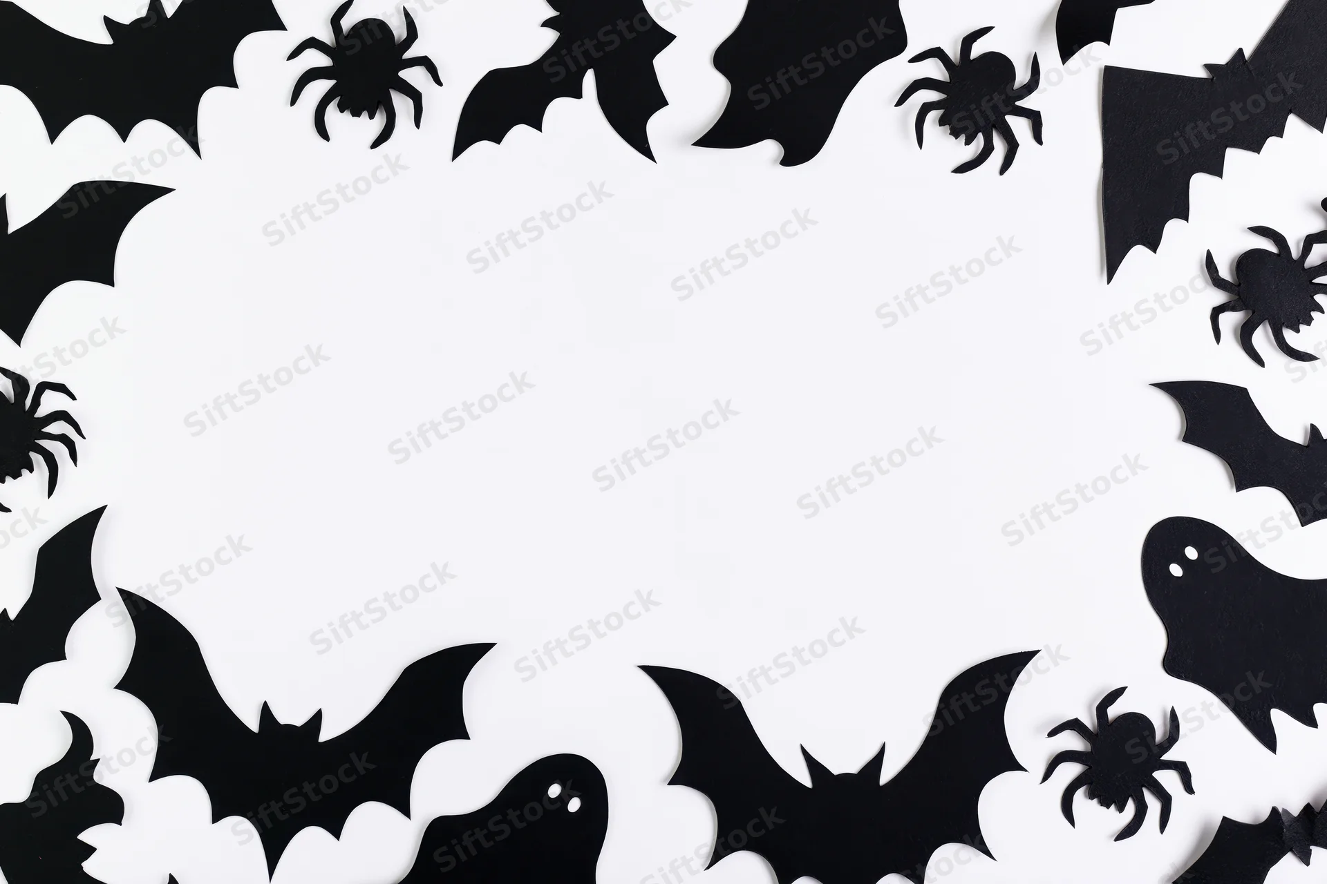 Black paper halloween cutouts of bats spiders and ghosts arranged in a frame on white background with copyspace