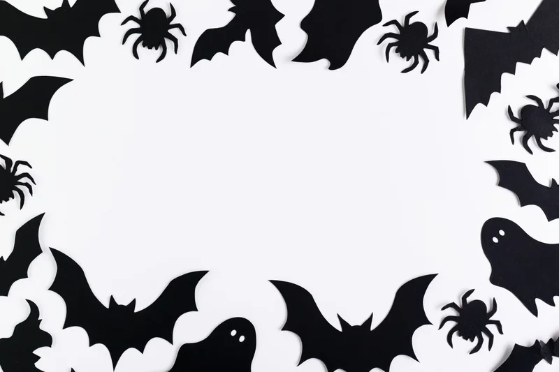 Black paper halloween cutouts of bats spiders and ghosts arranged in a frame on white background with copyspace