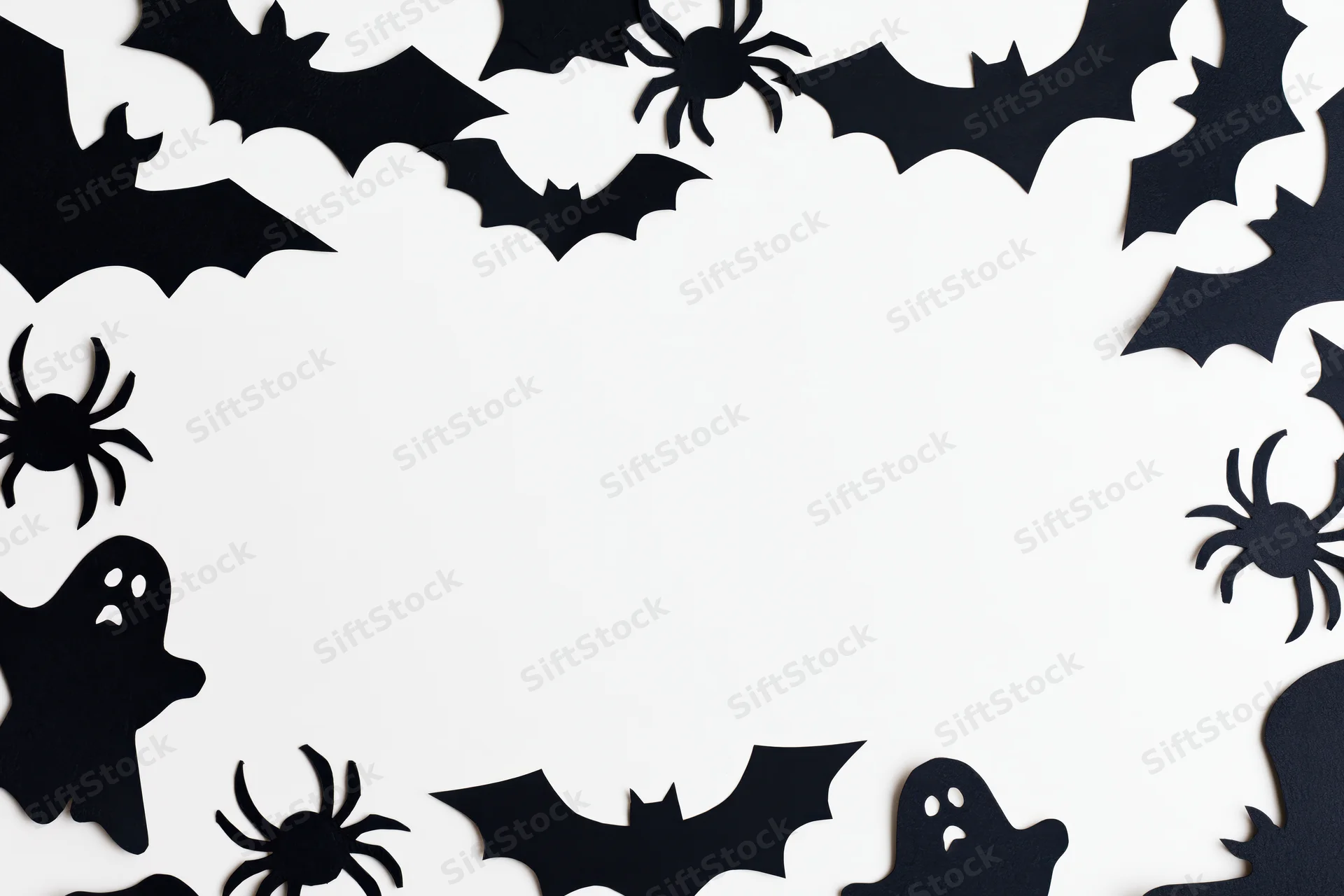 Black paper halloween cutouts of bats spiders and ghosts arranged in a frame on white background with copyspace