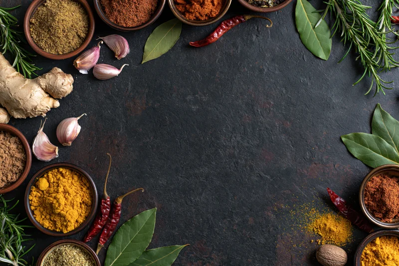 Cooking spices and herbs arranged in a border around a dark slate surface with copyspace in center