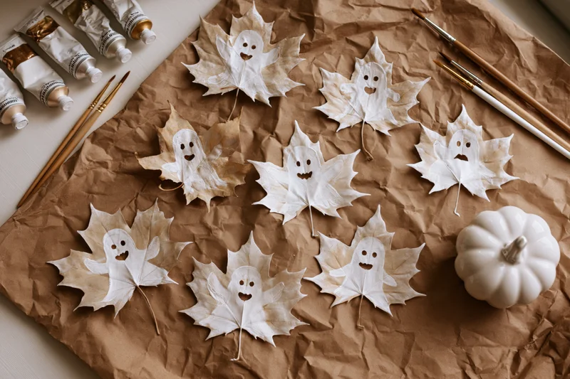 Dried maple leaves painted white with ghost faces arranged on brown paper with paint tubes and brushes for halloween craft