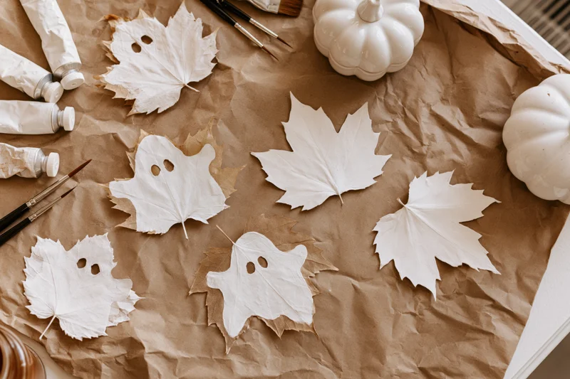 White-painted maple leaves shaped like ghosts arranged on crumpled brown paper with paint tubes and brushes for halloween craft