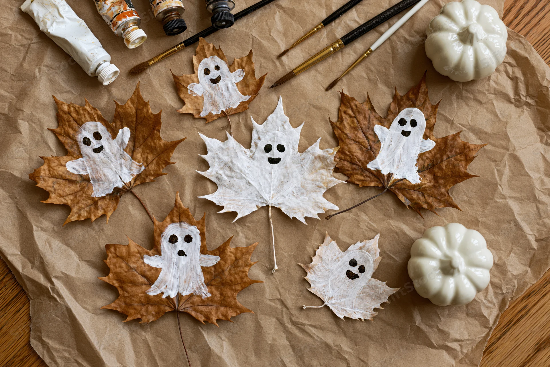 Dried autumn leaves painted as ghosts arranged with craft supplies and mini pumpkins on crumpled paper