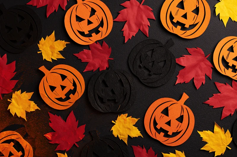 Handmade paper jack-o-lanterns and autumn leaves arranged in a flat lay on dark surface