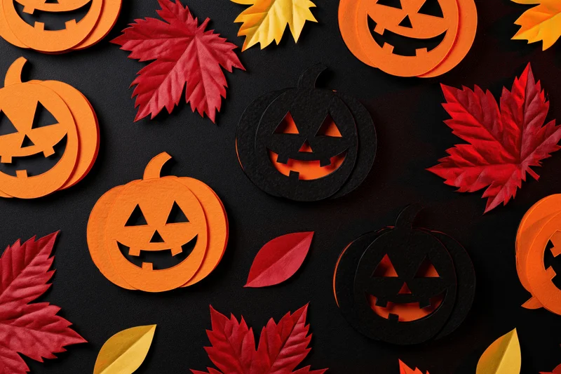 Handmade paper jack-o-lanterns and autumn leaves arranged in a flat lay on black background