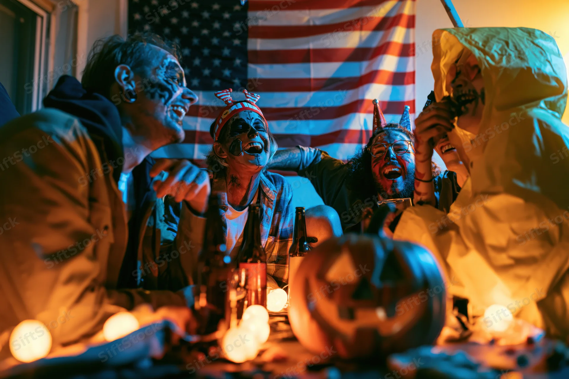 Four adults in halloween face paint and costumes laughing together at a decorated table with pumpkin and beer bottles indoors