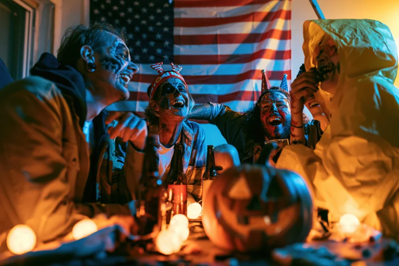 Four adults in halloween face paint and costumes laughing together at a decorated table with pumpkin and beer bottles indoors