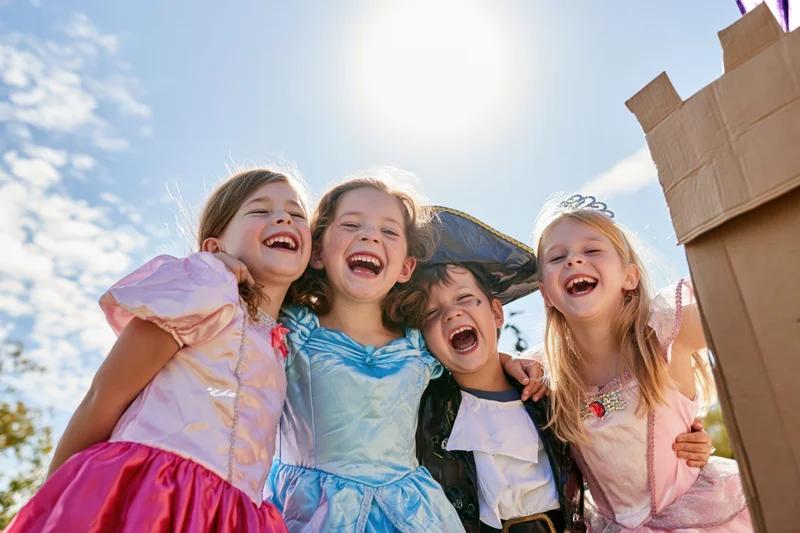 Four children in halloween costumes laughing together outdoors near a cardboard castle