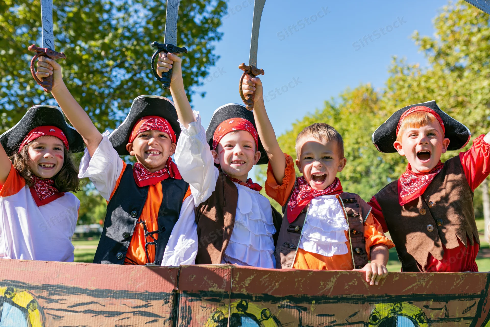 Children in pirate costumes holding swords and smiling behind a painted cardboard ship in a park