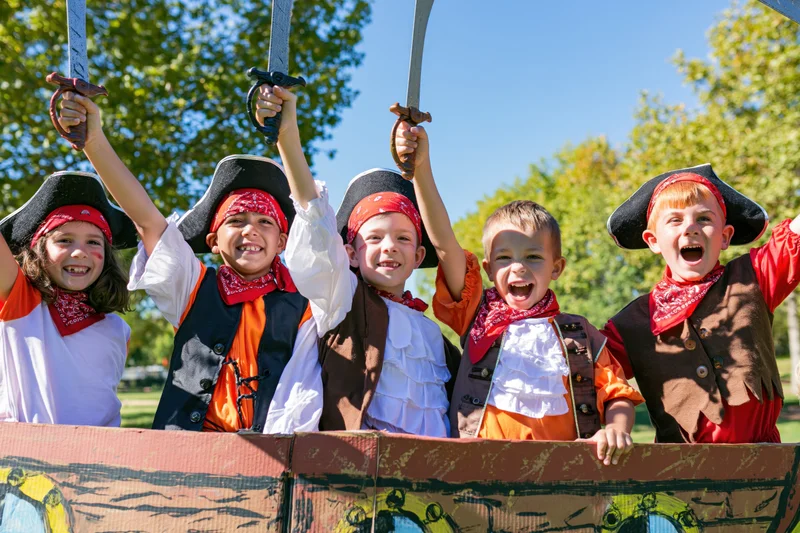 Children in pirate costumes holding swords and smiling behind a painted cardboard ship in a park
