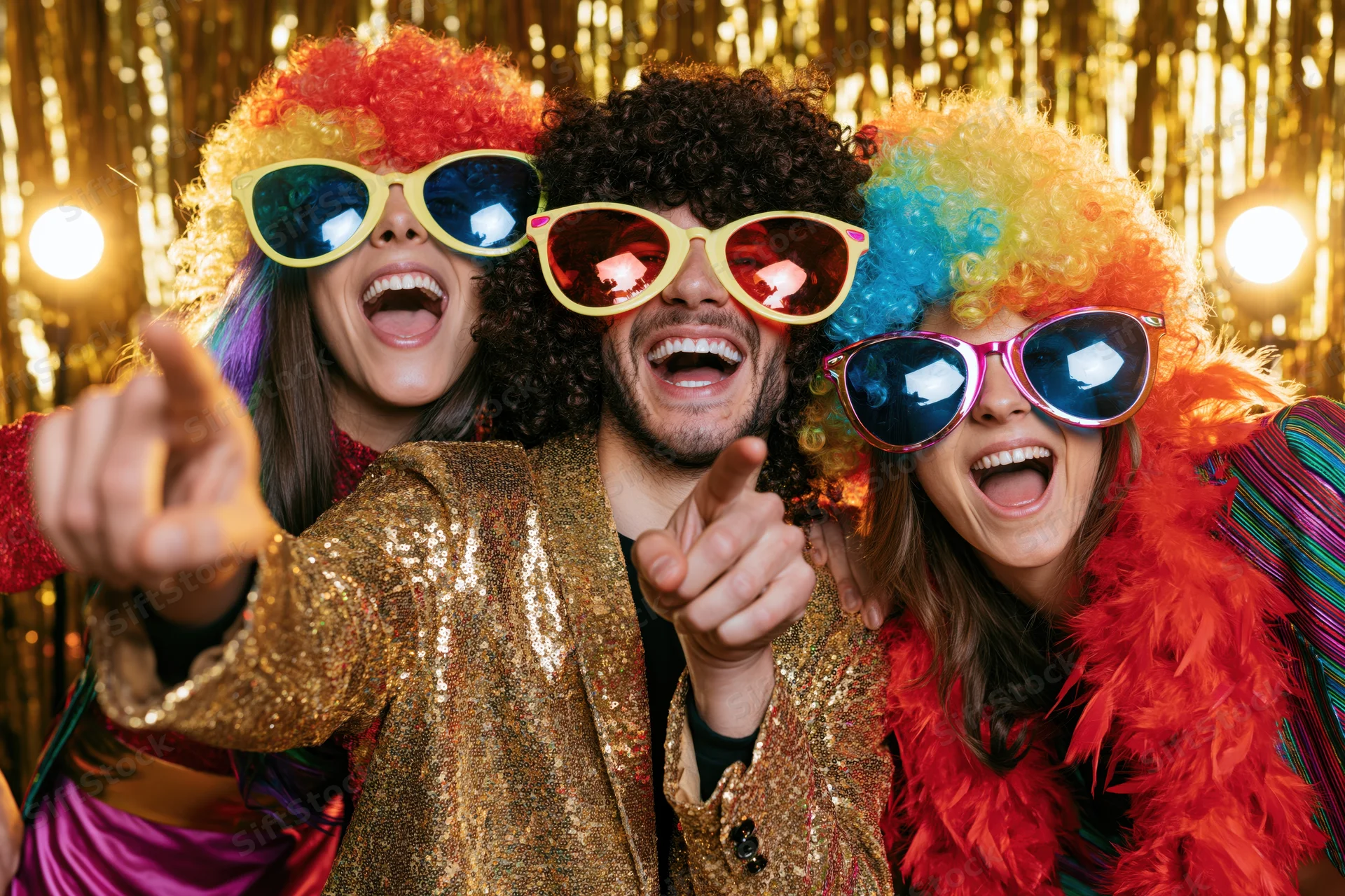 Three friends in retro disco costumes laughing and pointing at the camera in front of a gold sequin backdrop