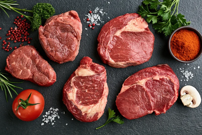 Raw beef steaks of different cuts arranged with herbs, spices, and vegetables on dark slate surface