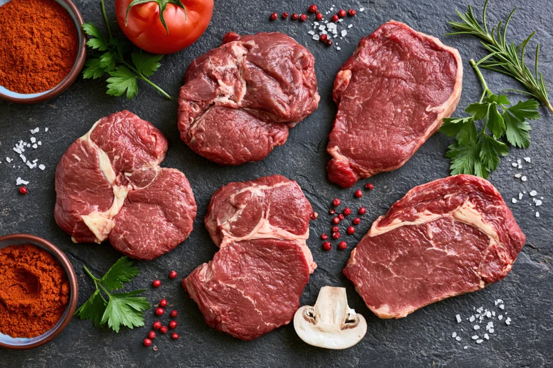 Raw beef steaks of different cuts arranged with herbs and spices on dark slate surface