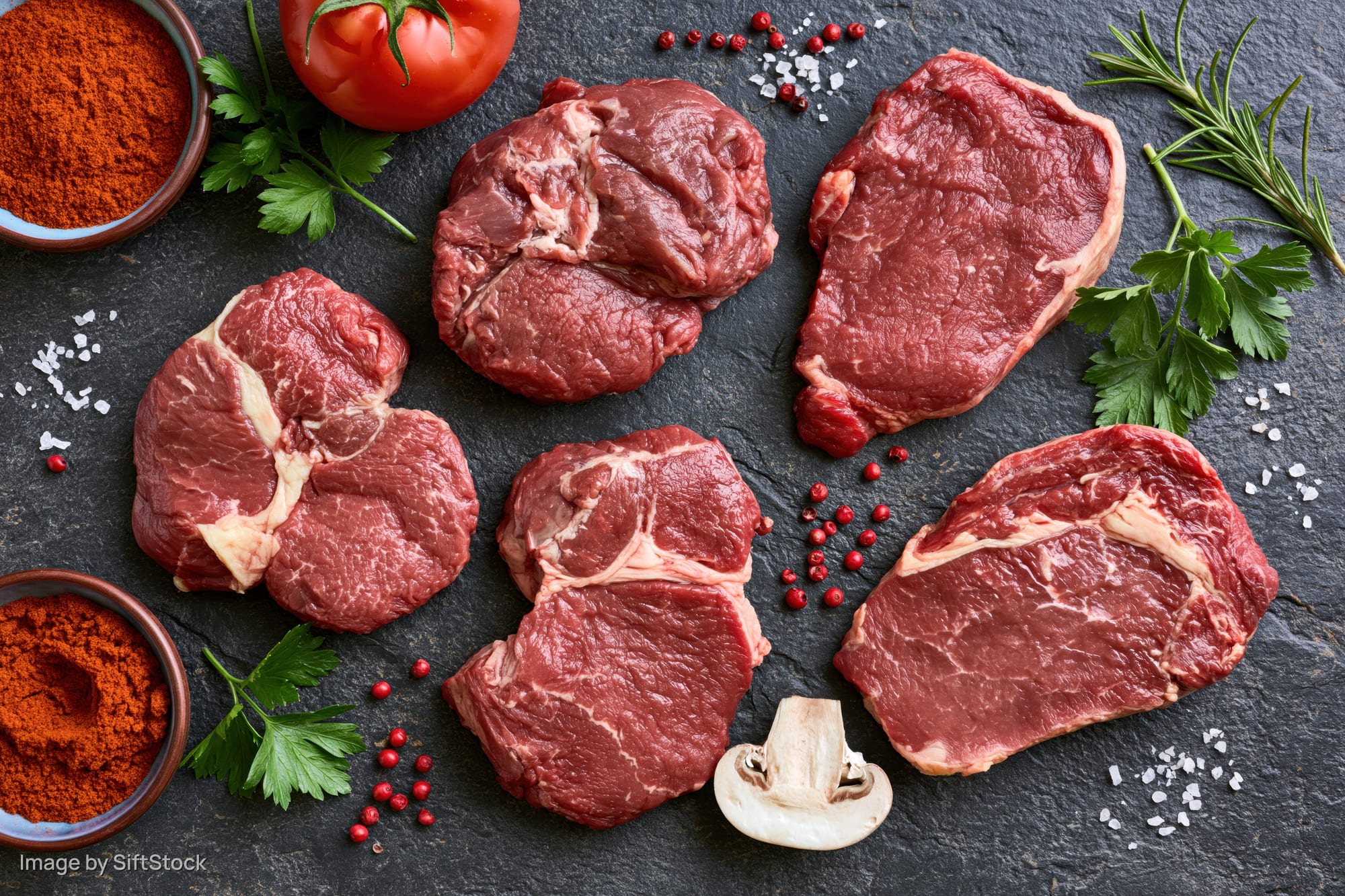 Raw beef steaks of different cuts arranged with herbs and spices on dark slate surface