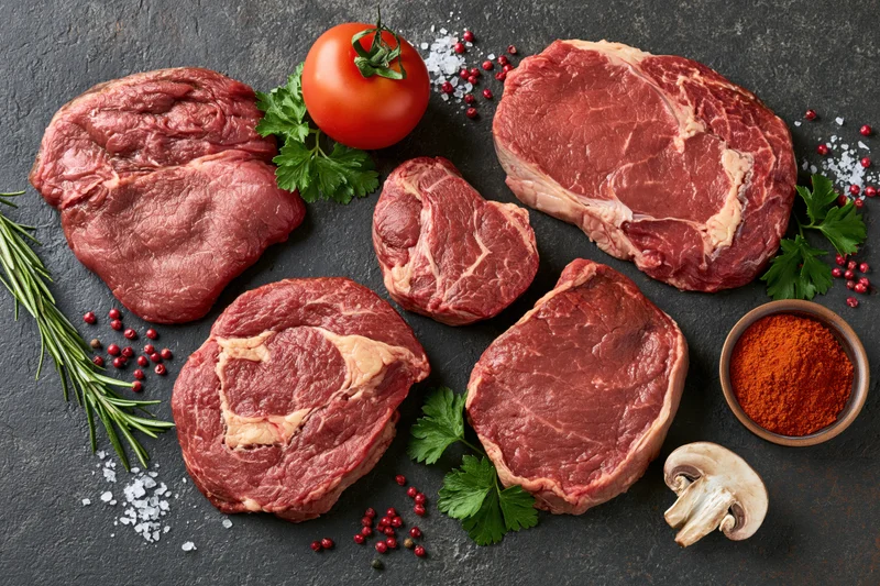 Raw beef steaks of different cuts arranged with herbs, spices, tomato and mushroom on dark surface