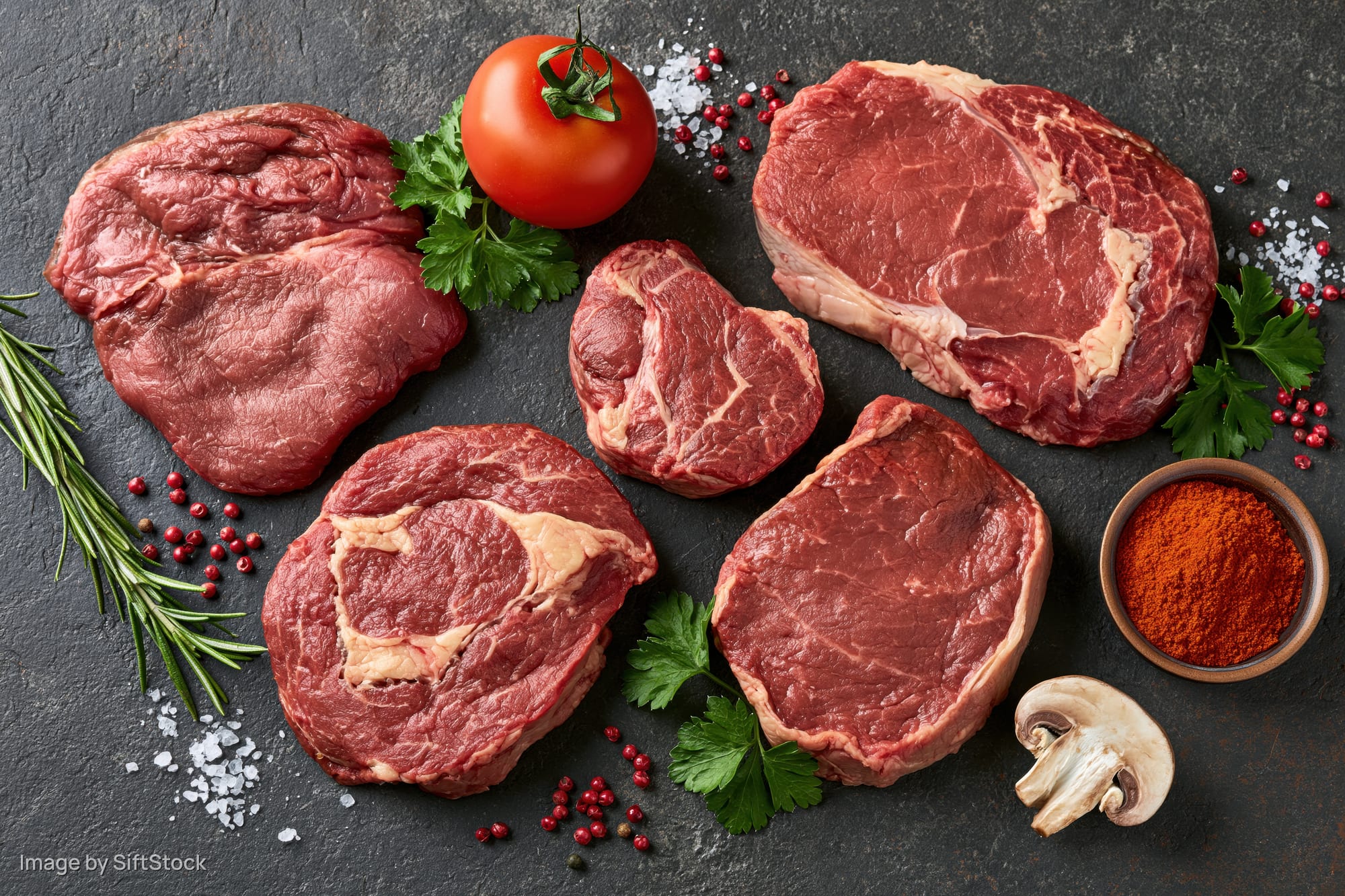 Raw beef steaks of different cuts arranged with herbs, spices, tomato and mushroom on dark surface