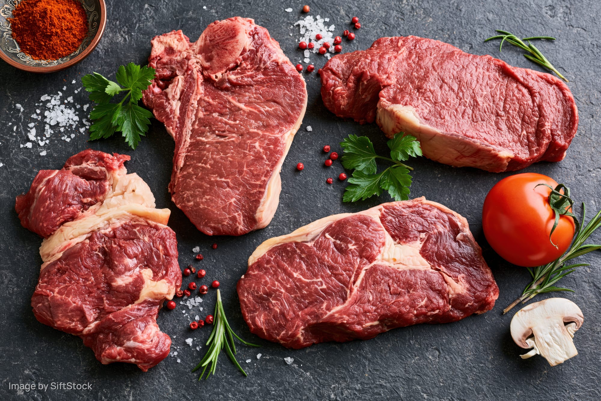 Four raw beef steaks of different cuts arranged on dark slate surface with herbs, tomato, mushroom, salt, pepper, and paprika garnish