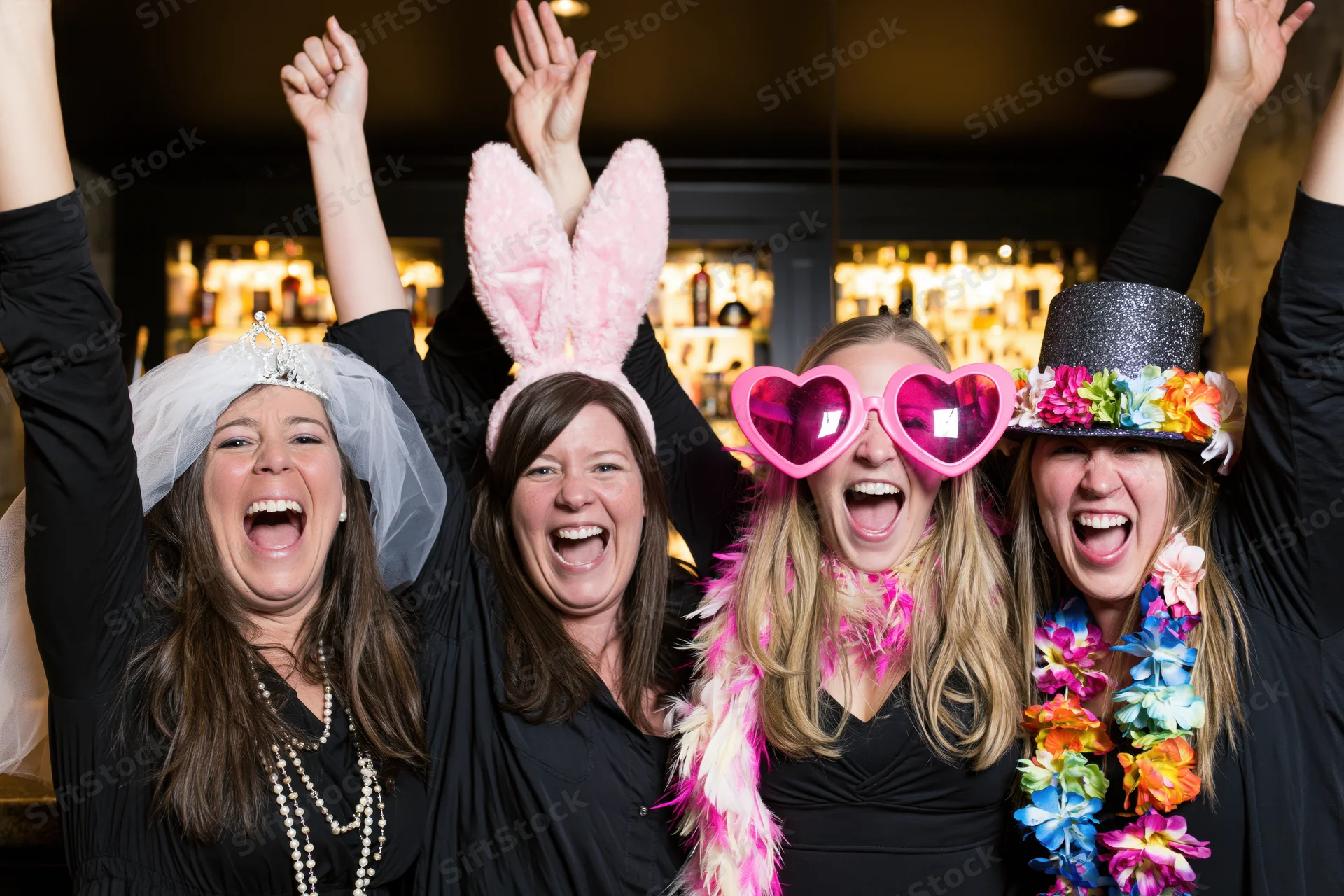 Four women laughing with arms raised at a nightclub party wearing festive accessories